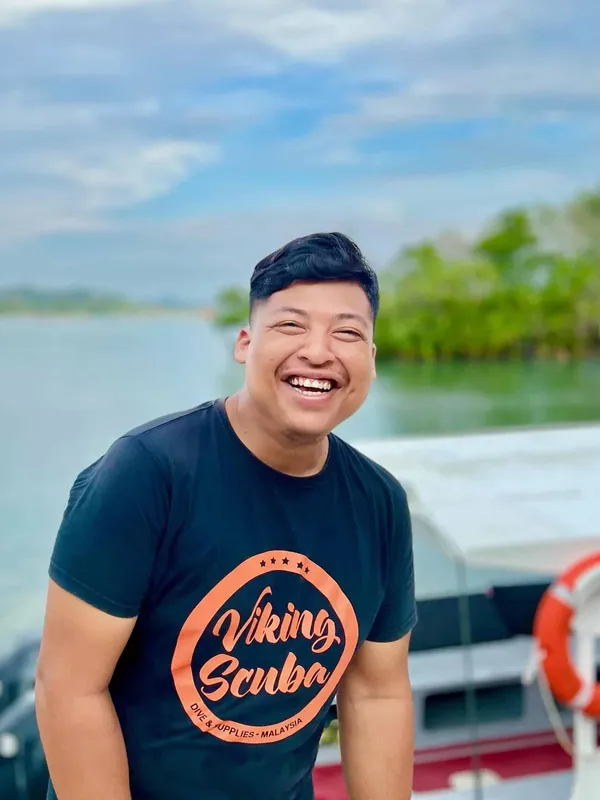 homme souriant portant un t-shirt noir devant un bateau et l'eau