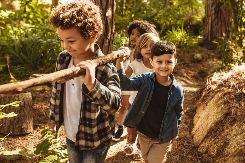 Quatre enfants souriants marchent en forêt, portant une branche