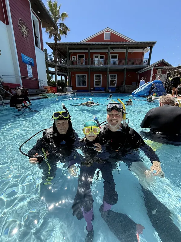 Trois plongeurs souriants en combinaison dans une piscine avec un bâtiment