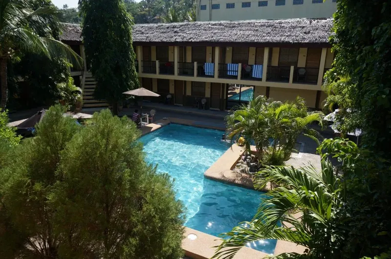 Bâtiment beige avec piscine et végétation tropicale, quelques personnes présentes