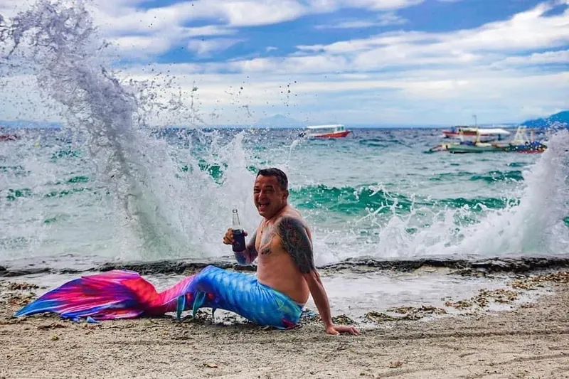 Homme déguisé en sirène sur une plage avec des vagues et bateaux