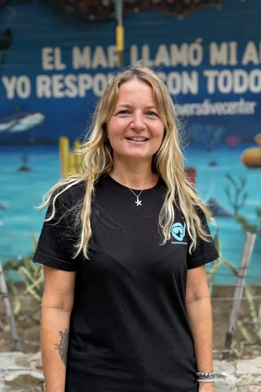 Femme souriante, cheveux blonds, portant un t-shirt noir devant un mur peint