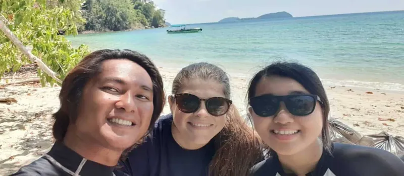 Trois personnes souriantes sur une plage paradisiaque avec un bateau en arrière-plan