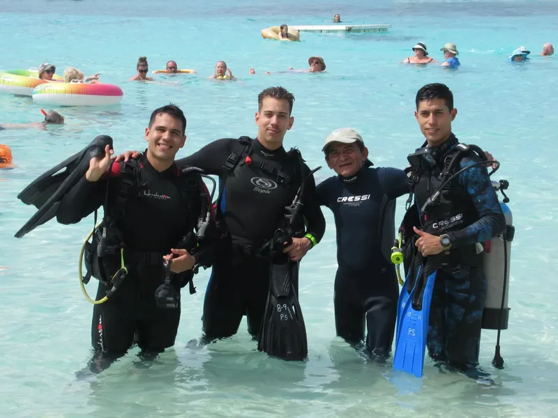 Quatre plongeurs souriants en combinaison, équipés, dans une eau turquoise claire