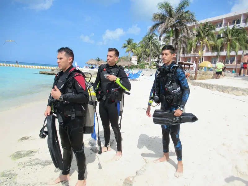 Trois plongeurs équipés sur une plage de sable blanc ensoleillée