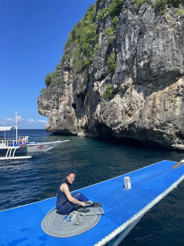Plongeuse souriante assise sur un bateau bleu devant une falaise