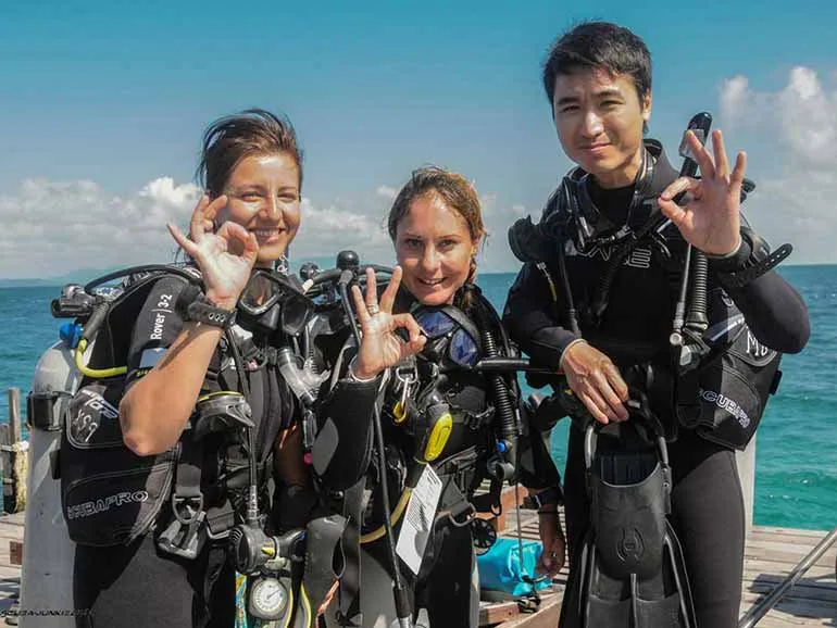 Trois plongeurs souriants font un signe de victoire sur un ponton