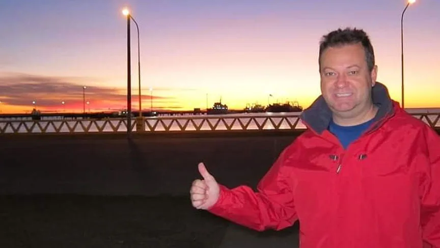 Homme souriant avec veste rouge, pouce levé, coucher de soleil en arrière-plan