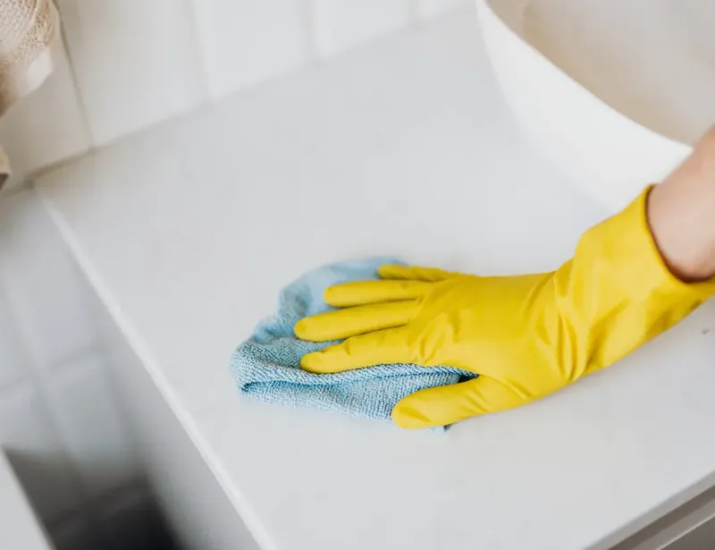 Main gantée jaune nettoyant une surface blanche avec un chiffon bleu