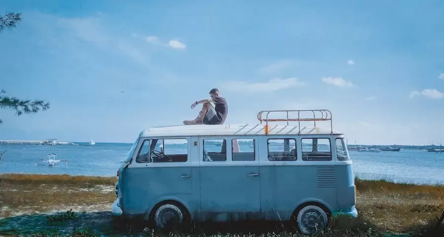 Un homme assis sur le toit d'un vieux combi bleu face à la mer