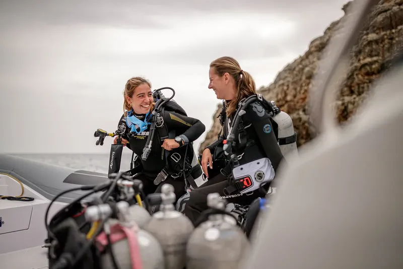 Deux plongeuses souriantes équipées discutent près d'un bateau et des bouteilles