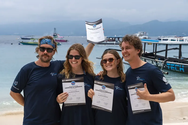 Quatre personnes souriantes présentent leurs certificats de plongée sur une plage
