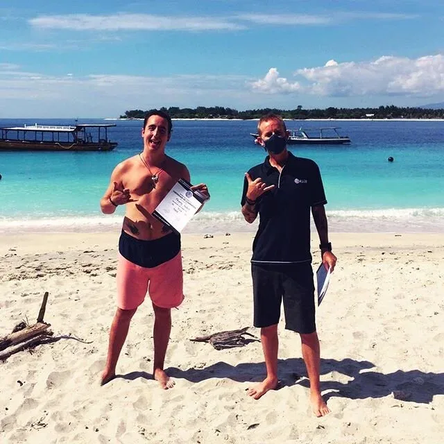 Deux hommes souriants sur une plage tropicale, tenant des certificats