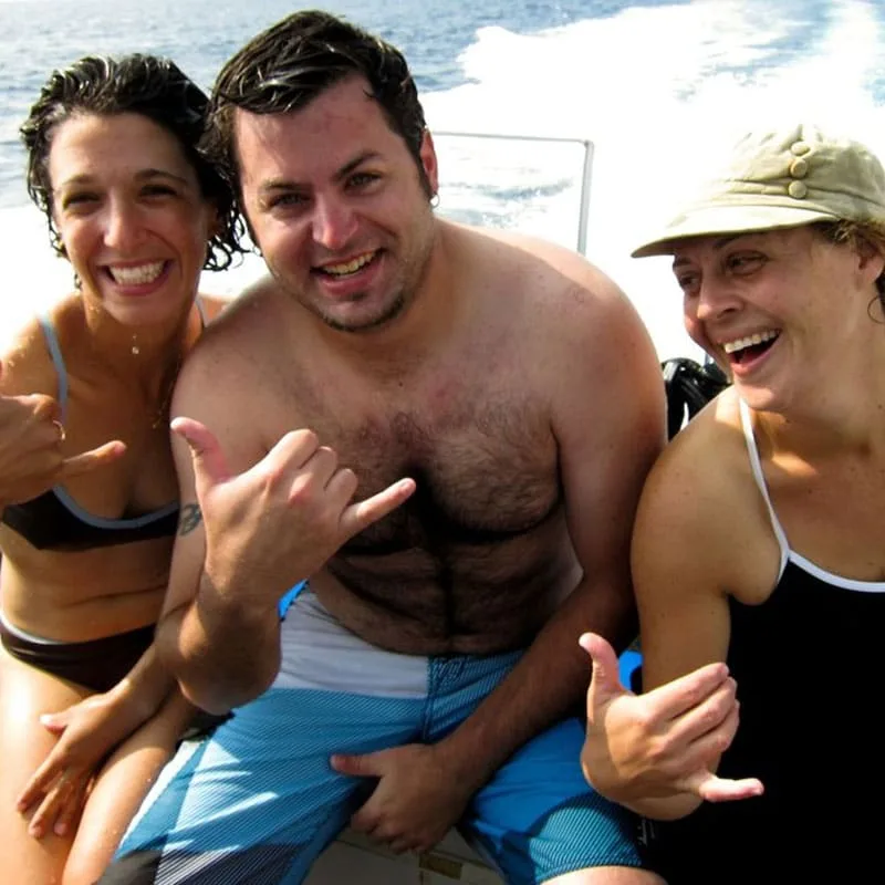 Trois personnes souriantes sur un bateau en mer, signe de la main