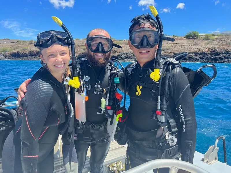 Trois plongeurs souriants en combinaison, masques et détendeurs sur un bateau