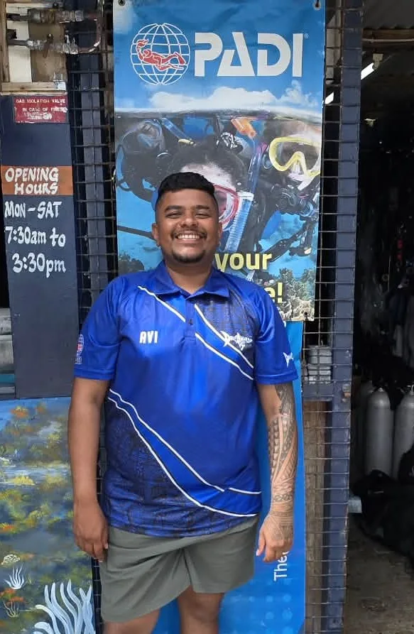 Un homme souriant pose devant une bannière PADI et du matériel de plongée