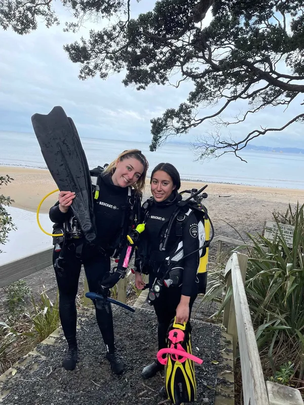 Deux plongeuses souriantes équipées près d'une rambarde en bois et d'une plage