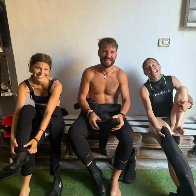 Trois personnes souriantes, en tenue de plongée, assises sur des palettes