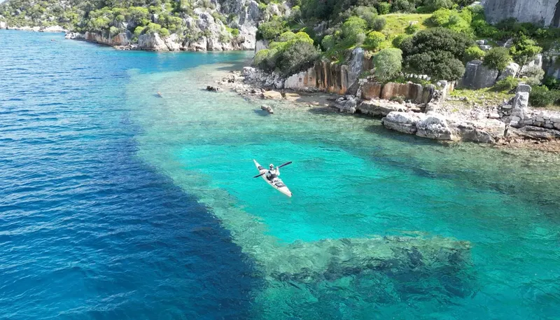 Kayak transparent sur eau turquoise, côte rocheuse et végétation luxuriante