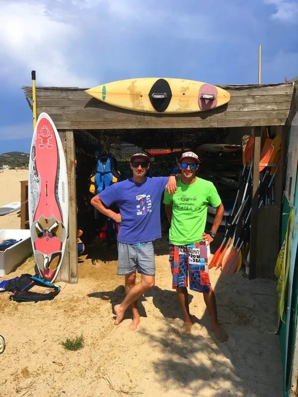Deux hommes souriants posent devant des planches de surf et du matériel
