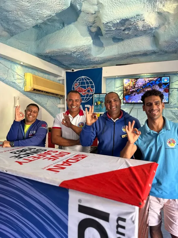 Quatre personnes souriantes, vêtues de bleu et turquoise, font signe