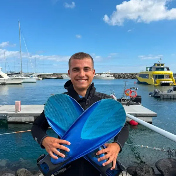 Un plongeur souriant tient des palmes bleues devant un port de plaisance