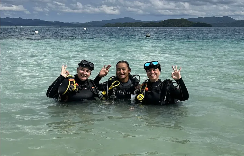 Trois plongeuses souriantes font signe dans une eau turquoise claire