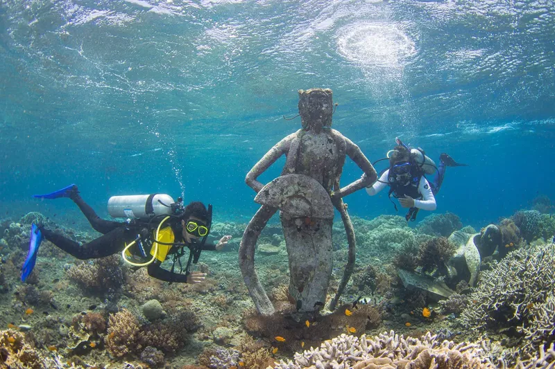 Deux plongeurs explorent une statue sous-marine au milieu des coraux