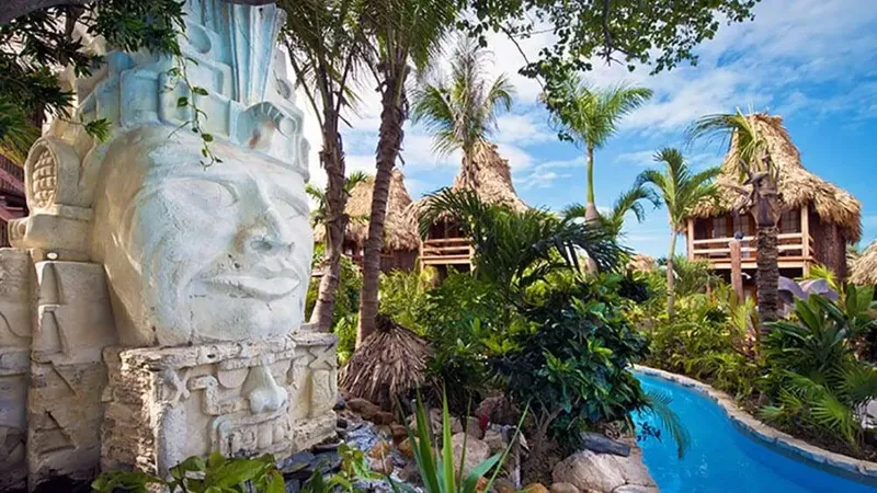 Statue de visage maya, végétation tropicale, cabanes sur pilotis, ciel bleu