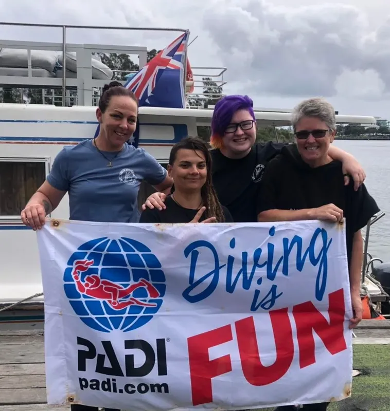 Quatre personnes souriantes tiennent une bannière PADI devant un bateau