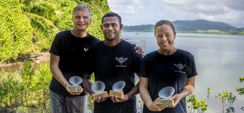 Trois personnes souriantes tiennent des trophées devant un paysage marin
