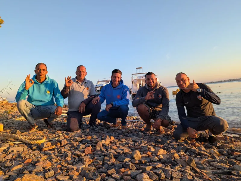 Cinq personnes souriantes accroupies sur une plage de galets au lever du soleil
