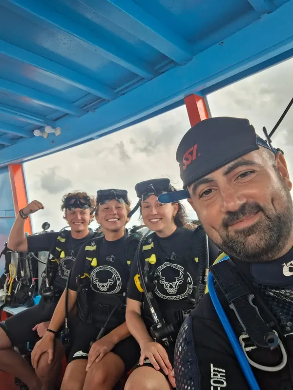 Quatre plongeurs souriants équipés sur un bateau, prêts à plonger
