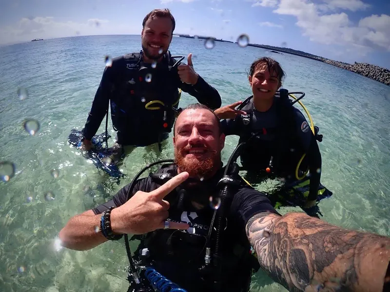 Trois plongeurs souriants en combinaison, eau turquoise, ciel bleu clair