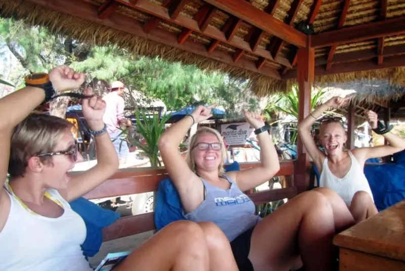 Quatre plongeuses souriantes, bras levés, sous un abri en chaume