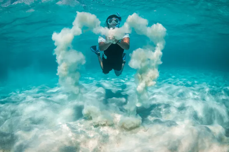 Plongeur en combinaison noire entouré de sable et de bulles sous-marines