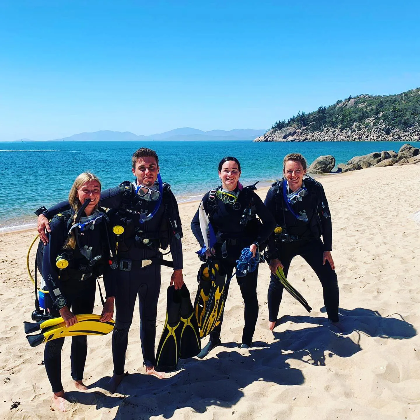 Quatre plongeurs souriants en combinaison sur une plage ensoleillée
