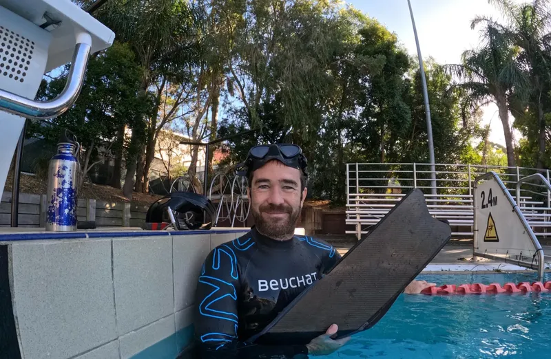 Plongeur souriant avec masque, combinaison et palme devant une piscine