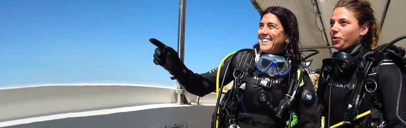 Deux plongeuses souriantes sur un bateau, équipées et pointant au loin
