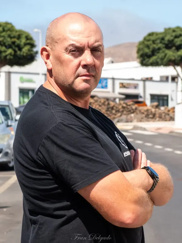 Homme chauve regardant la caméra, bras croisés, devant un bâtiment