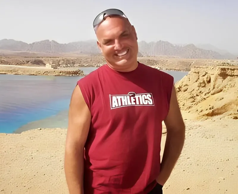 Homme souriant en t-shirt rouge, paysage montagneux et mer en arrière-plan