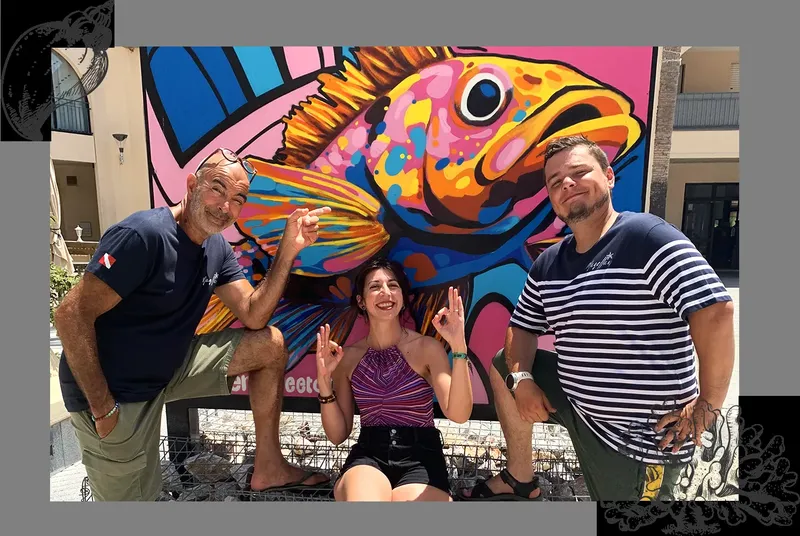 Trois personnes souriantes posent devant une fresque de poisson coloré