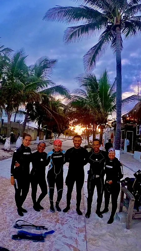 Groupe de plongeurs en combinaison, souriants, devant un bâtiment tropical