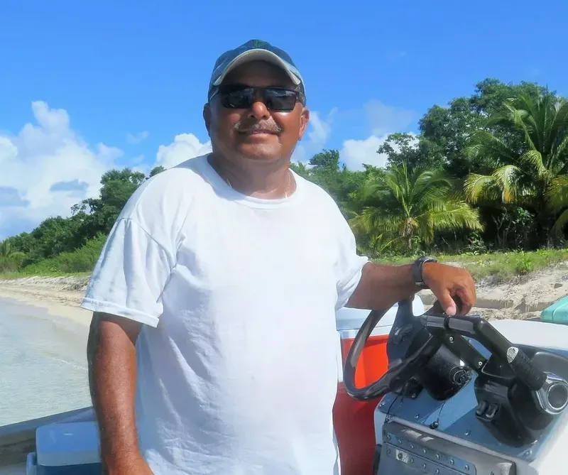 Homme souriant sur un bateau, devant une plage et végétation tropicale