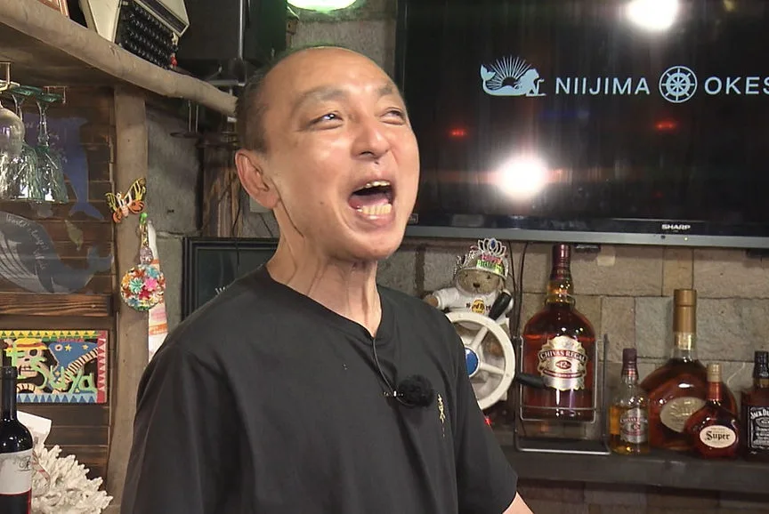 Homme asiatique souriant devant une étagère de bouteilles et un écran