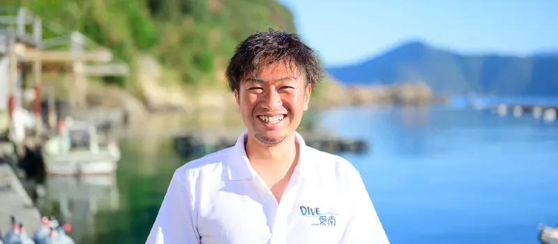 Homme souriant sur un port avec des bateaux et montagnes en arrière-plan