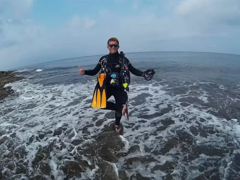 Plongeur en combinaison, palmes et masque, souriant sur une plage rocheuse