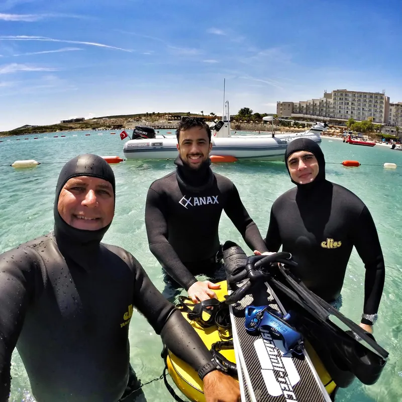 Trois plongeurs en combinaison sourient devant un bateau et la mer