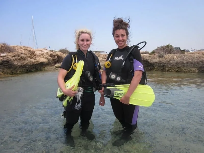 Deux plongeuses souriantes en combinaison et palmes dans l'eau peu profonde