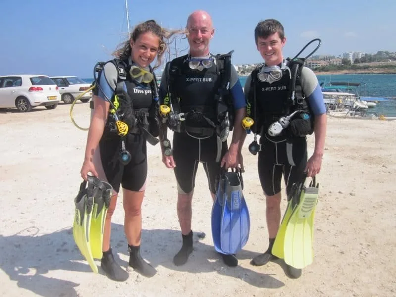 Trois plongeurs équipés sur une plage de sable avec des bateaux en arrière-plan